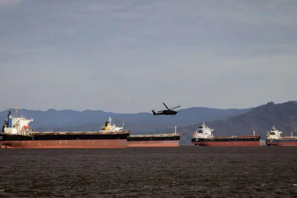 A helicopter flies over several cargo ships in a vast ocean with a mountainous backdrop.