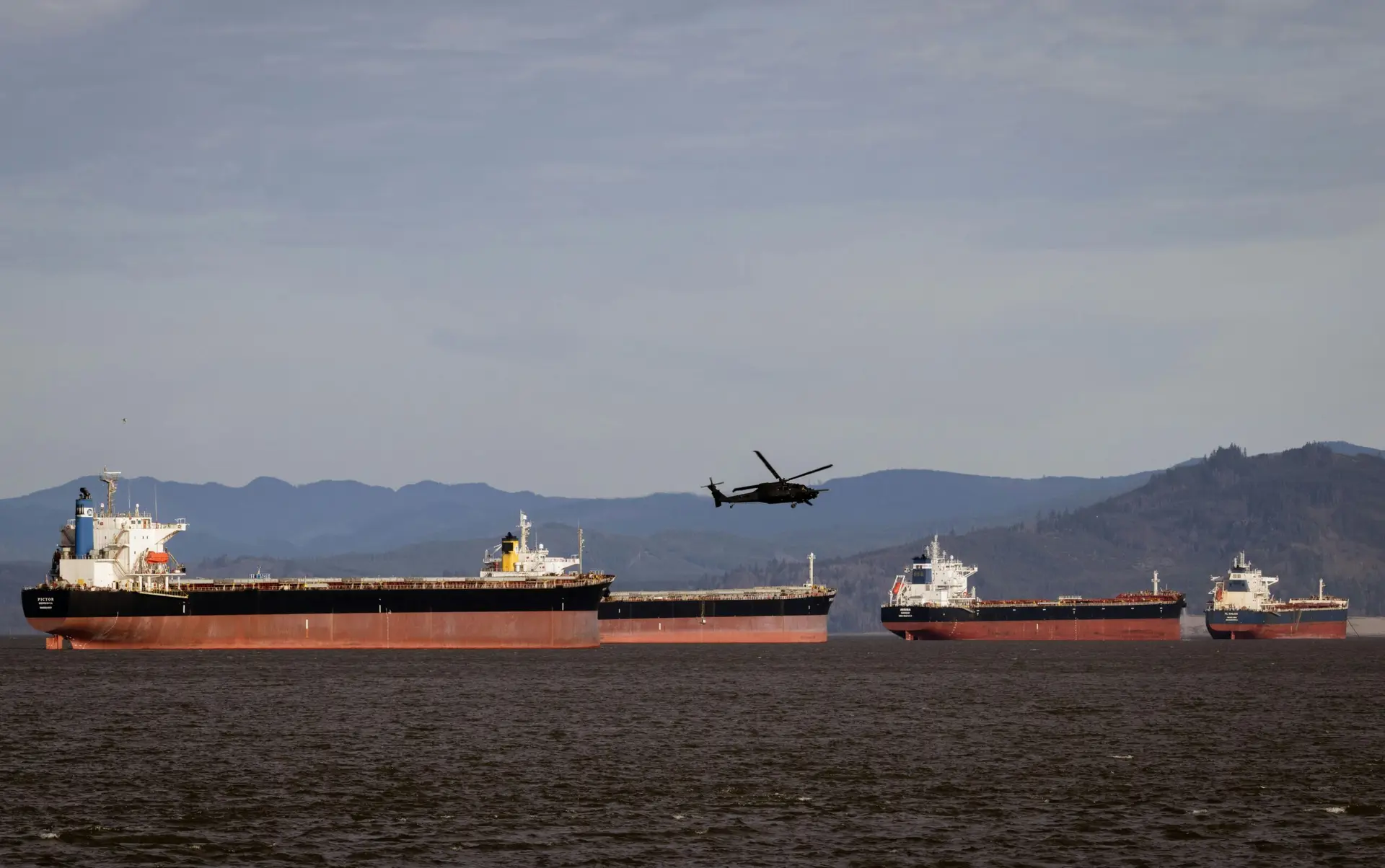A helicopter flies over several cargo ships in a vast ocean with a mountainous backdrop.